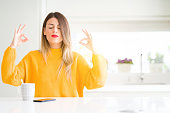 Young beautiful woman drinking a cup of coffee at home relax and smiling with eyes closed doing meditation gesture with fingers. Yoga concept.