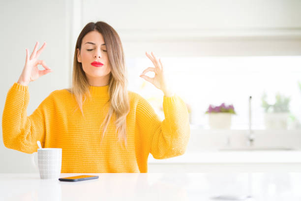 jovem e bela mulher bebendo uma xícara de café em casa relaxar e sorrir com os olhos fechados, fazendo o gesto de meditação com os dedos. conceito de ioga. - smiling serene people yoga women - fotografias e filmes do acervo