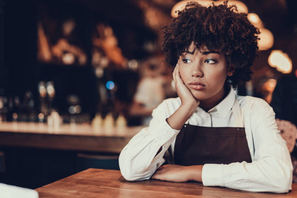 Tired young lady looking aside and sitting over table Portrait of upset waitress. She looking aside and sitting behind table while touching cheek by palm hand waitress stock pictures, royalty-free photos & images