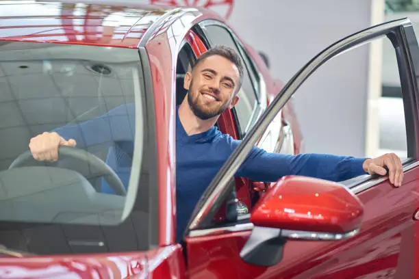 Young handsome man almost sitting in red car and closing front door. Brunette client in car salon smiling from car and holding hand on steering wheel. Happy driver testing new vehicle before buying.