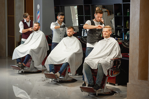 Three barbers in white shirts and waistcoat doing styling of hair and beard with scissors and straight razor in barber shop. Young handsome men sitting in chair during procedure. Concept of haircut.