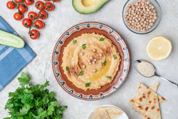 istock Hummus in a brown clay plate with a blue pattern. On the white table are vegetables, greens, triangular pieces of pita. Top view. Flat lay. 1097843654