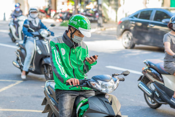 Grab motorbike taxi in Vietnam Da Nang, Vietnam - October 14, 2018: A young man on scooter, wearing a Grab green helmet and jacket, looks at his mobile phone. Grab app is a popular way of earning money as well as traveling. vietnamese man taxi motorbike driver stock pictures, royalty-free photos & images