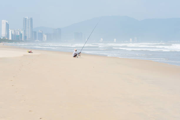 Danang, Vietnam - November 1, 2018: a man fishes with a spinning rod in the shallow wavy sea waters sitting on the sunlit empty beach with Da Nang city skyline in the background.