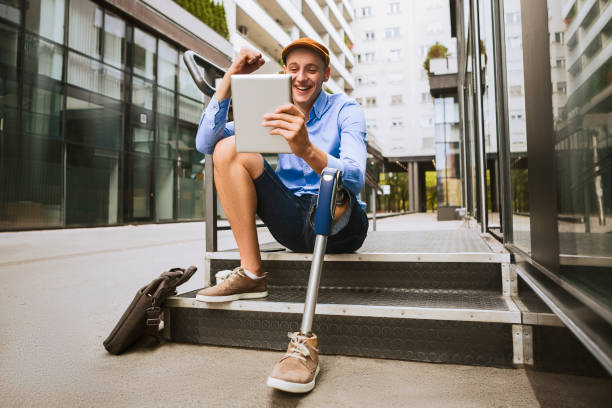 mann mit beinprothese sitzen vor das bürogebäude während des gesprächs auf einem digitalen tablet - people equipment healthcare and medicine slim stock-fotos und bilder