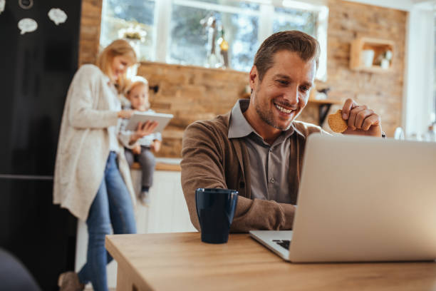 Working from home to be close to his family Cropped shot of a young man working on the laptop with his family in the background Petrovaradin stock pictures, royalty-free photos & images