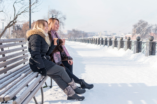 Two laughing friends sitting on a bench with a cup of coffee at the winter promenade