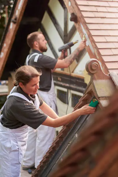 a male and female painter team set about painting a mock tudor style house from scaffold. The female painter is holding a sanding block and smiling to camera.