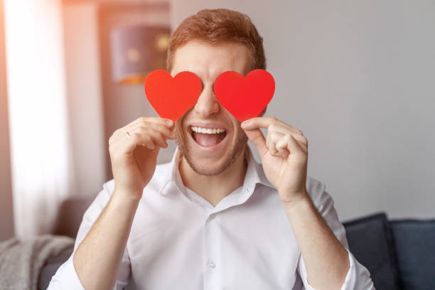 Handsome male keeping two red hearts near eyes while celebrating Valentines Day at home