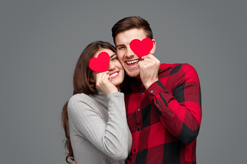 Excited young man and woman smiling and hugging each other while standing on gray background and holding tiny hearts near eyes