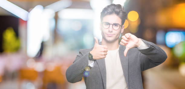 young business man wearing glasses over isolated background doing thumbs up and down, disagreement and agreement expression. crazy conflict - thumbs up human thumb human hand conflict imagens e fotografias de stock