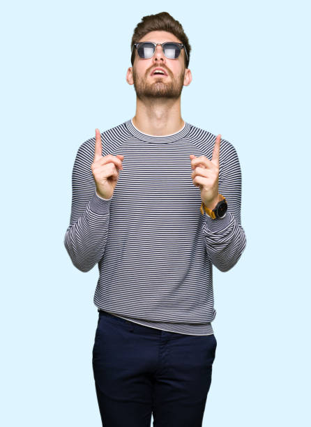 istock Young handsome man wearing sunglasses amazed and surprised looking up and pointing with fingers and raised arms. 1097997068