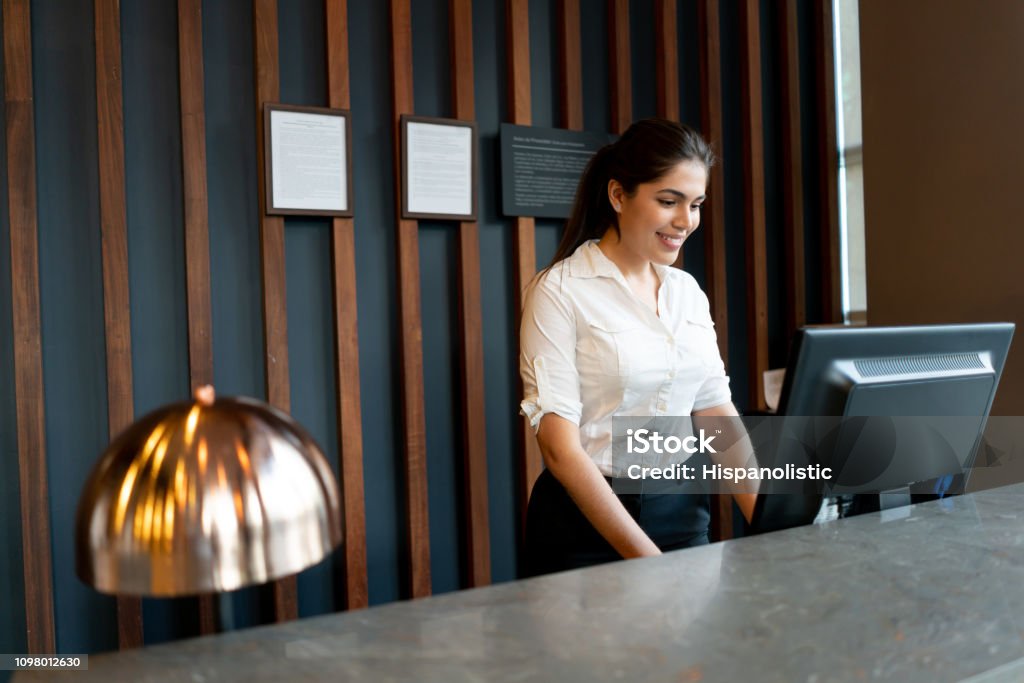 Latin american hotel receptionist working behind counter smiling while looking at computer screen Latin american hotel receptionist working behind counter smiling very happy while looking at computer screen Hotel Stock Photo