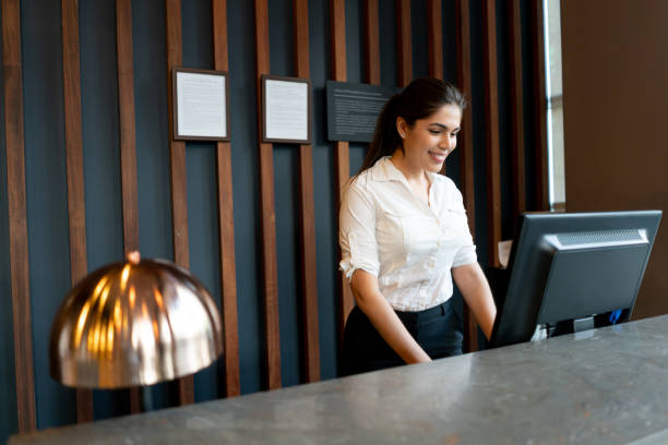Latin american hotel receptionist working behind counter smiling while looking at computer screen Latin american hotel receptionist working behind counter smiling very happy while looking at computer screen concierge photos stock pictures, royalty-free photos & images