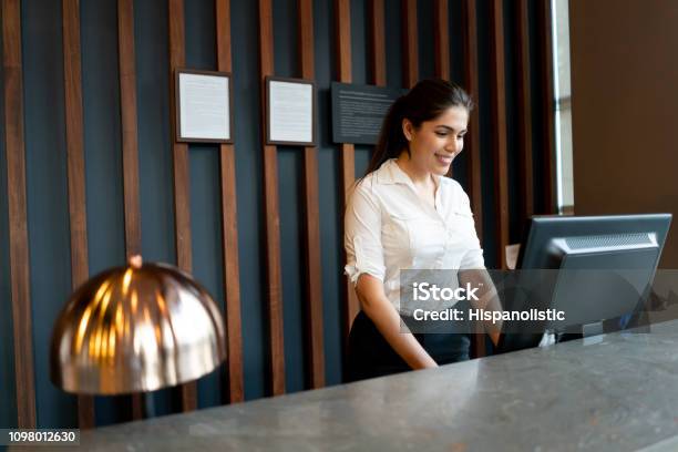 Latin American Hotel Receptionist Working Behind Counter Smiling While Looking At Computer Screen Stock Photo - Download Image Now
