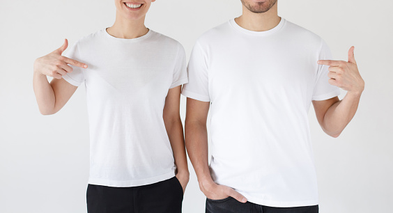 Smiling young couple pointing at blank white t-shirts with index fingers, copy space for your advertising, isolated on gray background