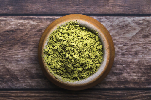 Close-up of a bowl filled with green matcha tea powder. The brown ceramic bowl is on an old dark wooden table.  The angle of the photo is straight from above.