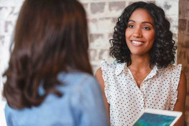 Business colleagues having a conversation. Business colleagues having a conversation. They are both young business people casually dressed in a modern office. Could be an interview or consultant working with a client. She is listening and smiling. One person has her back to us. She is holding a digital tablet. Mixed ethnic group.  One is African American and the other is Caucasian casual job interview stock pictures, royalty-free photos & images