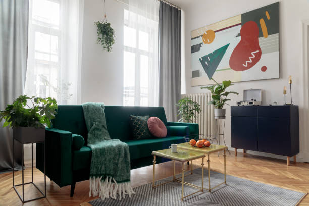 istock Sunny and bright luxury home interior with design velvet sofa, armchair, tables, pouf, commode and accessroies. White walls with painting. Sunny view. Stylish decor of sitting room. Big windows. 1098075036