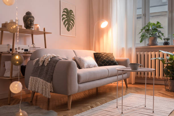 istock Stylish scandinavian interior with design sofa, poster, plants, pillows, bookstand, coffee table, cozy blanket and mock up frames. White background walls, brown wooden parquet. Photo by night. 1098075640