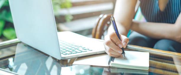 Business woman hand is writing on a notepad with a pen and using a laptop computer. stock photo