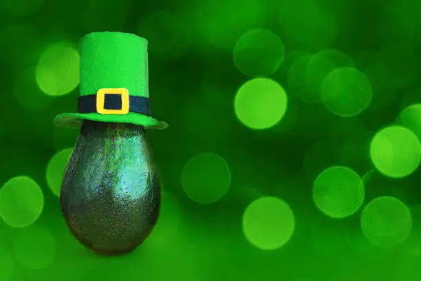 St. Patrick's day decoration background idea with fresh green avocado in leprechaun hat on bright bokeh glittery background. Card concept. Closeup. Front view. Copy space