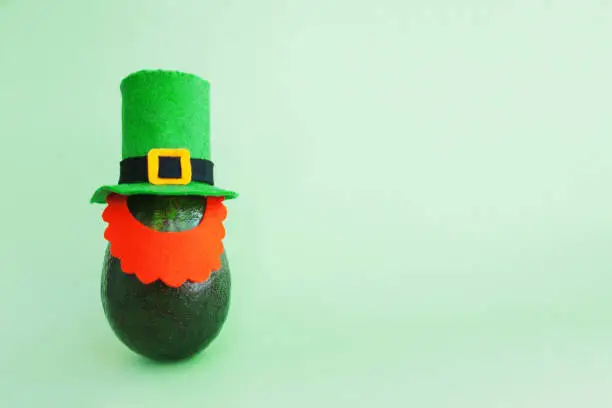 St. Patrick's day decoration background idea with fresh green avocado in leprechaun hat with a red beard of felt on bright background. Card concept. Closeup. Front view. Copy space