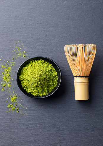 Matcha, green tea powder in black bowl with bamboo whisk on slate background. Top view