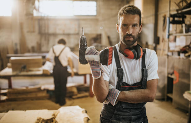 Portrait of manual worker with a drill in carpentry workshop. Young carpenter holding a drill and looking at camera while standing in a workshop. there are people in the background. holding drill stock pictures, royalty-free photos & images