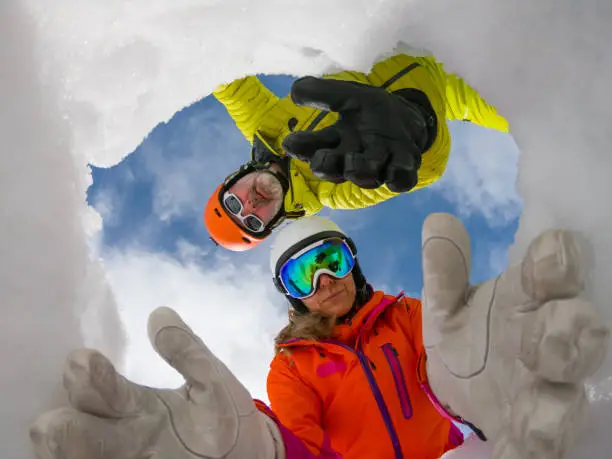 burried fisheye camera in snow hole - concept shot for avalanche rescue. Male and female skier in colorful winter clothes acting as montain rescue-service workers