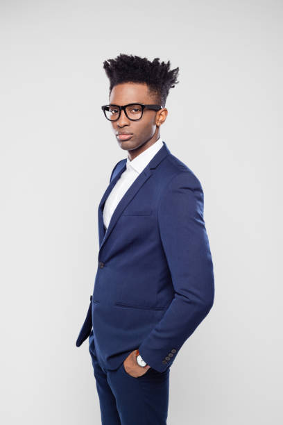 Portrait of handsome young afro american businessman standing with his hands in pocket on gray background. Man in business suit wearing eyeglasses looking at camera in studio.