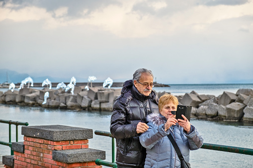 Salerno, Italy - Jnuary 03, 2019: A mature man and woman on the promenade in Salerno, Italy looking at the display on an electronic device during sunset. In the background a breakwater with Christmas lights, part of the yearly Luci d'Artista, festival of light.