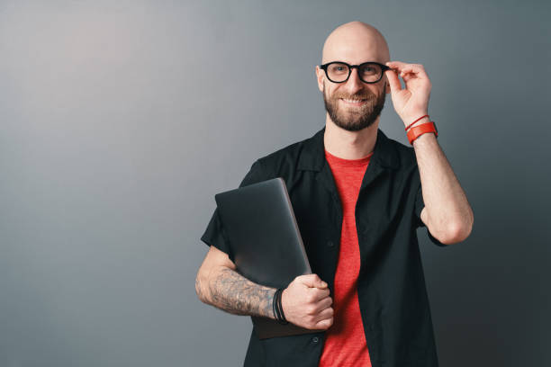 uomo barbuto sorridente con gli occhiali che tengono il laptop sotto il braccio toccando gli occhiali con le dita in studio su sfondo grigio - t shirt men red portrait foto e immagini stock