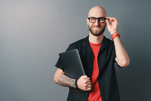 Smiling young bearded man holding laptop under the arm touching  his glasses with fingers, in the studio on gray background. Stylish designer, offer a best design experience to his clients.