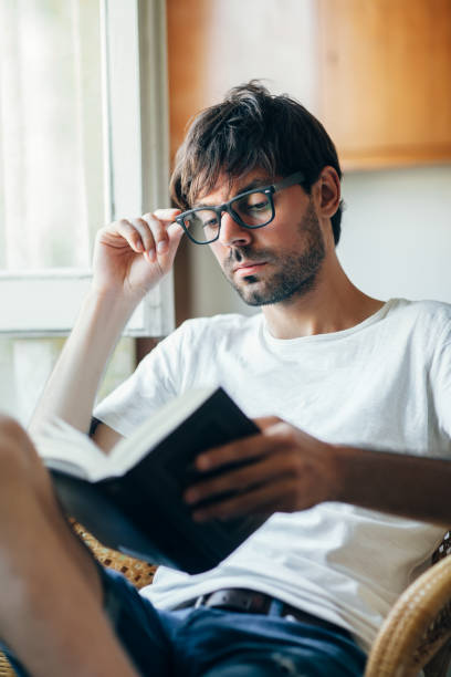 istock Young man in eyeglasses reading book sitting on chair 1098266488