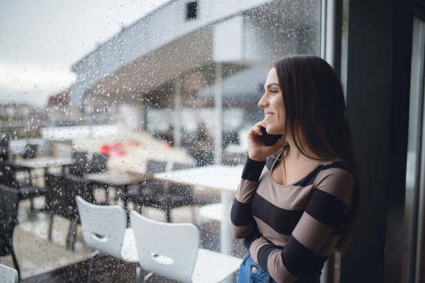 Looking through window Young woman posing by panoramic window and using phone rainy cafe stock pictures, royalty-free photos & images