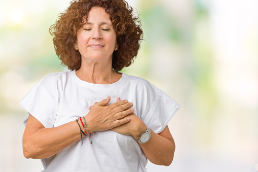 Beautiful middle ager senior woman wearing white t-shirt over isolated background smiling with hands on chest with closed eyes and grateful gesture on face. Health concept.