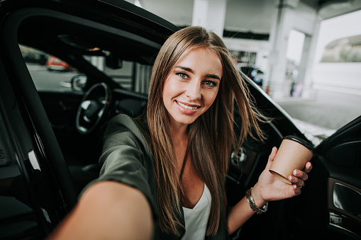 Portrait of happy female taking selfie while drinking mug of appetizing liquid during rest in contemporary vehicle