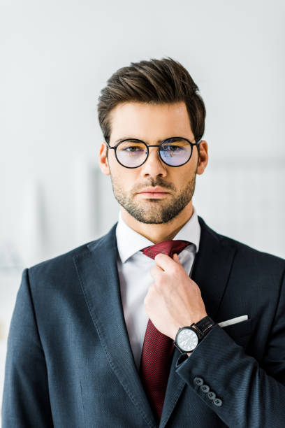 istock handsome businessman in formal wear and glasses adjusting tie while looking at camera in office 1098298074