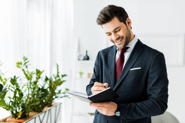 istock handsome smiling businessman in formal wear writing in notebook in office 1098298078