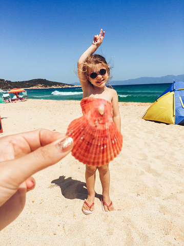Little girl standing at the beach while her mom making her dress of seashell