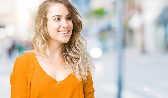 Beautiful young blonde woman over isolated background looking away to side with smile on face, natural expression. Laughing confident.