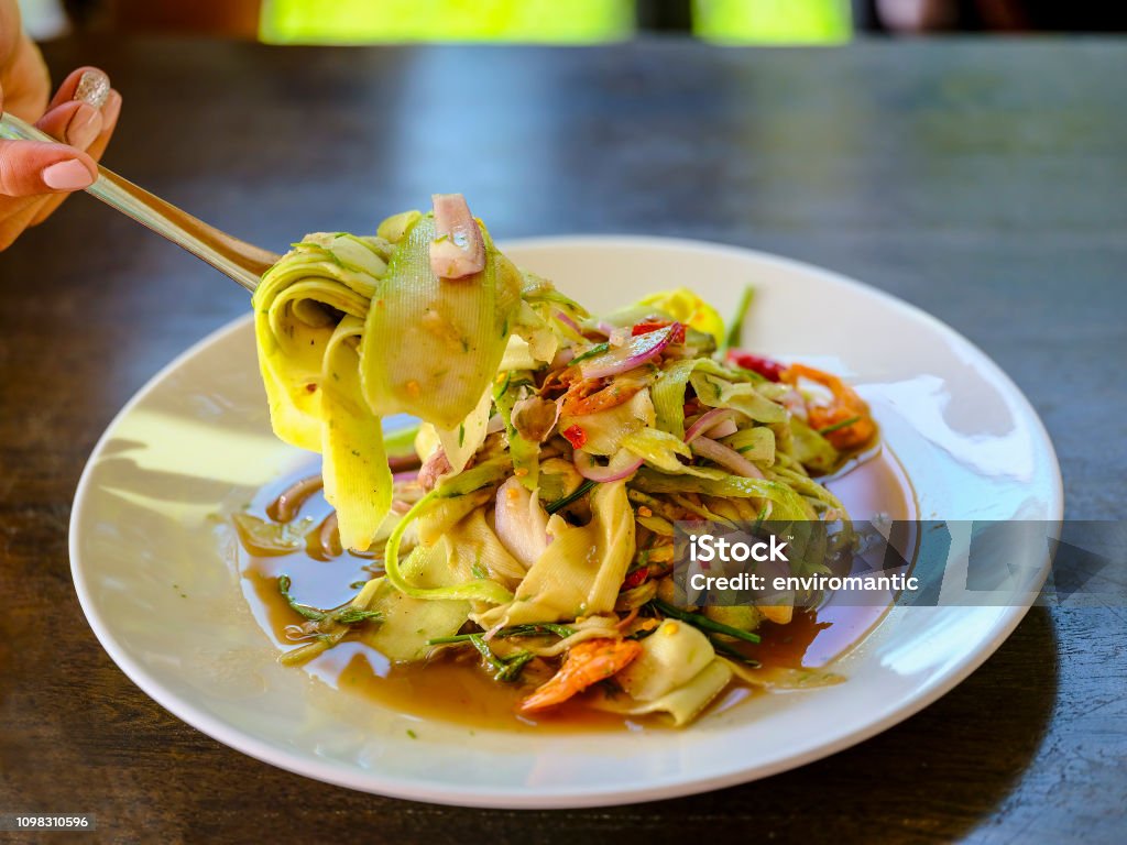 Thai woman using wooden chopsticks to eat a fresh delicious Thai spicy bamboo and prawn Thai style salad, Thailand. Adult Stock Photo