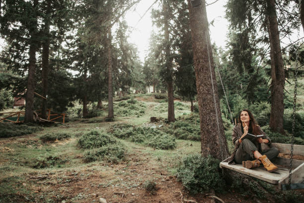 Charming girl holding notebook while sitting on wooden swing Peace and quiet. Portrait of beautiful young woman searching inspiration in forest. She is touching chin with pen and looking away with smile timberland arizona stock pictures, royalty-free photos & images