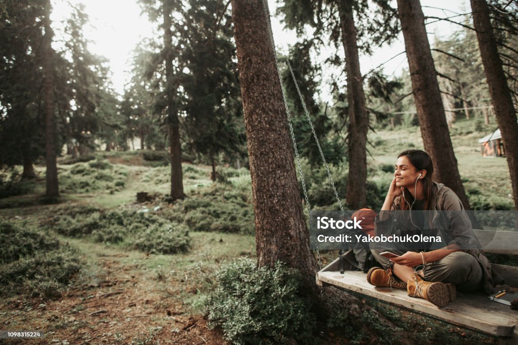 Young attractive woman listening music on smartphone in the forest In the woods. Portrait of beautiful girl with closed eyes sitting on wooden swing and enjoying favorite song. She is smiling Nature Stock Photo