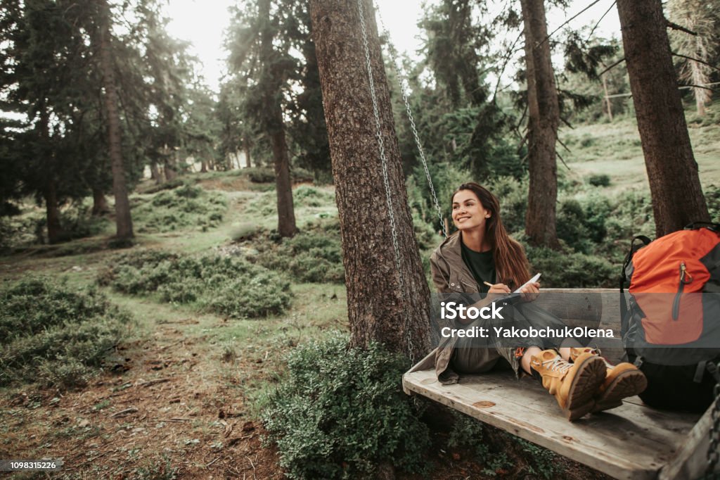 Beautiful girl making notes while sitting on wooden swing in forest I like this place. Portrait of charming young lady holding notebook with pen while looking away with smile. Trees and green plants on background Adult Stock Photo