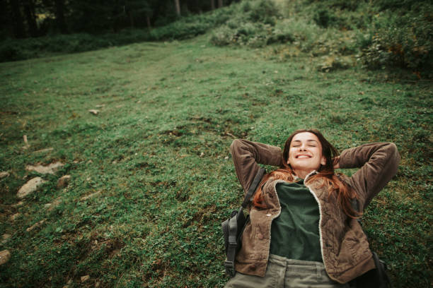 Smiling girl lying on back with hands above her head Fun trip. Portrait of beautiful girl with closed eyes resting on the grass during travelling in the forest women lying down grass wood stock pictures, royalty-free photos & images