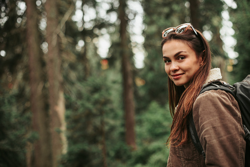 Enjoying hike. Portrait of smiling girl with backpack travelling alone. Coniferous wood on blurred background