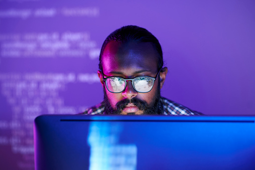 A focused man with a beard and glasses is working intently at a computer, surrounded by purple lighting and code displayed on a screen in the background.