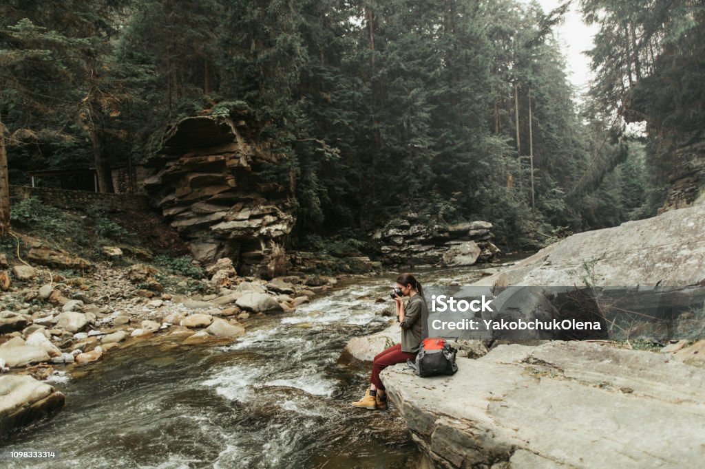 Pleasant female traveler making photos of natural beauty Nice young woman making photos while sitting on the bank of the river in the forest Adult Stock Photo